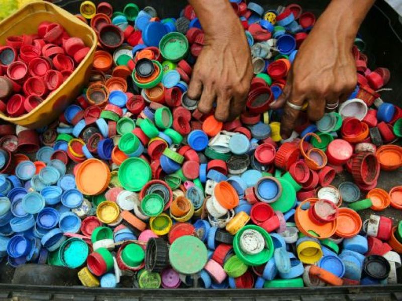 Un par de manos separa y organiza una gran cantidad de tapas plásticas de diferentes colores dentro de un contenedor. Esta actividad promueve el reciclaje y la reutilización de materiales, destacando la importancia de la gestión adecuada de residuos plásticos.